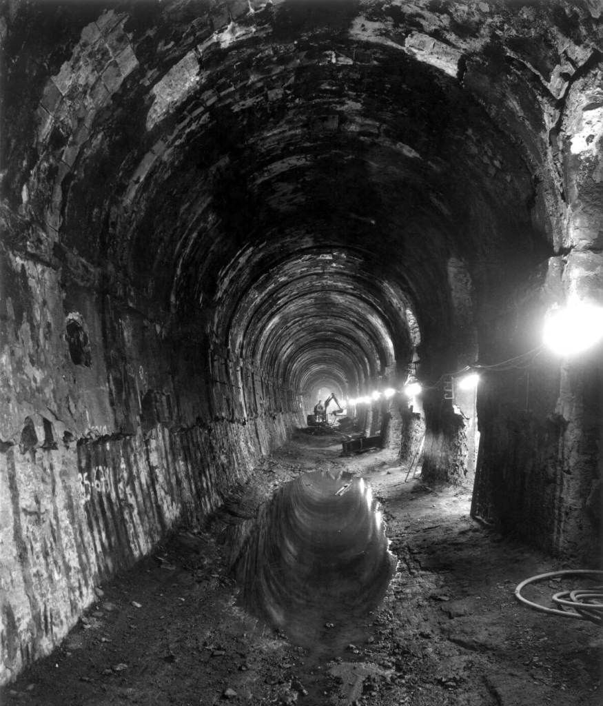 A black and white photograph of the interior of a large tunnel with a person sitting on a small digger in the background.