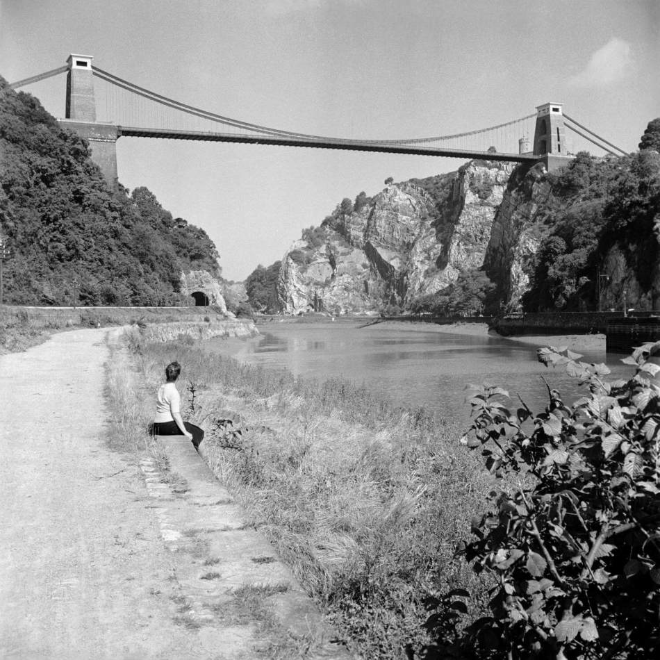 A black and white photograph a person sitting beside a river looking up at Clifton Suspension Bridge.