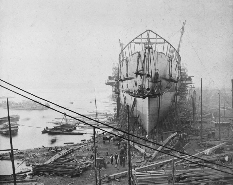 A black and white photograph of a large ship under construction in a dock.