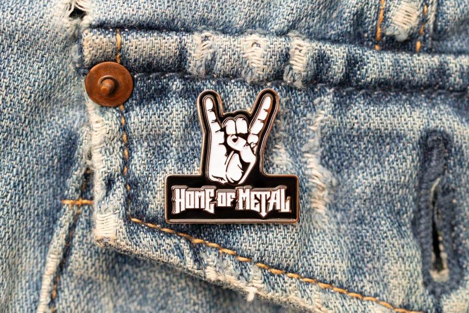 A photograph of a close-up of an enamel pin badge in the shape of a hand in a rock symbol with the wording 'Home of Metal' on it.