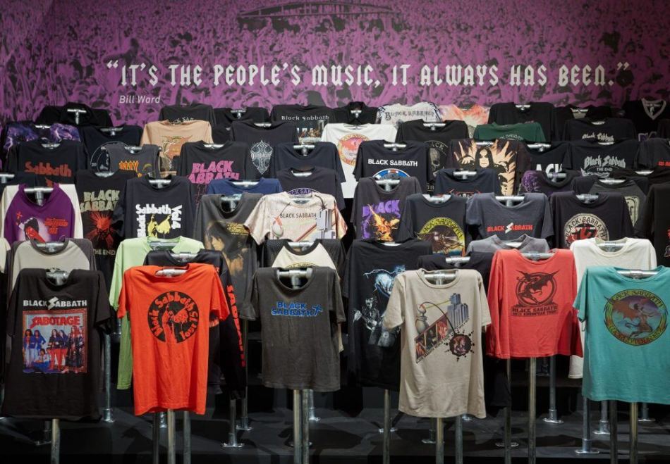 A photograph of a collection of Black Sabbath t-shirts displayed in an exhibition. Above them a sign reads: 'IT'S THE PEOPLE'S MUSIC, IT ALWAYS HAS BEEN - Bill Ward'.