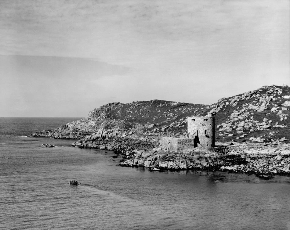 A black and white photograph of a small castle tower on the edge of a cliff beside the sea.