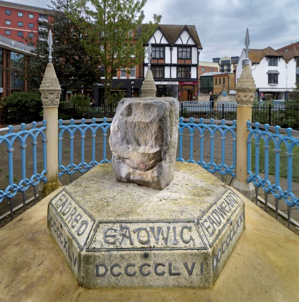 A photograph of a large stone on a plinth in the middle of a pathway surrounded by an ornate iron fence.