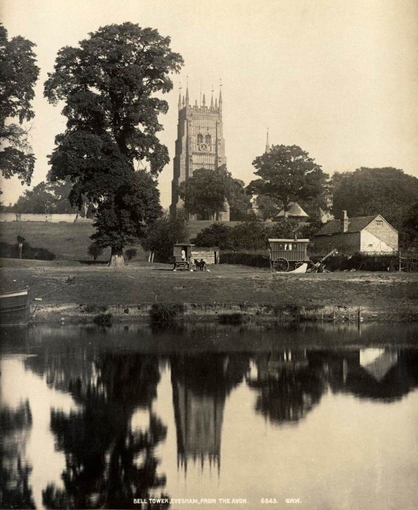 A black and white photograph of a view across a river with horse-drawn caravans in the background.