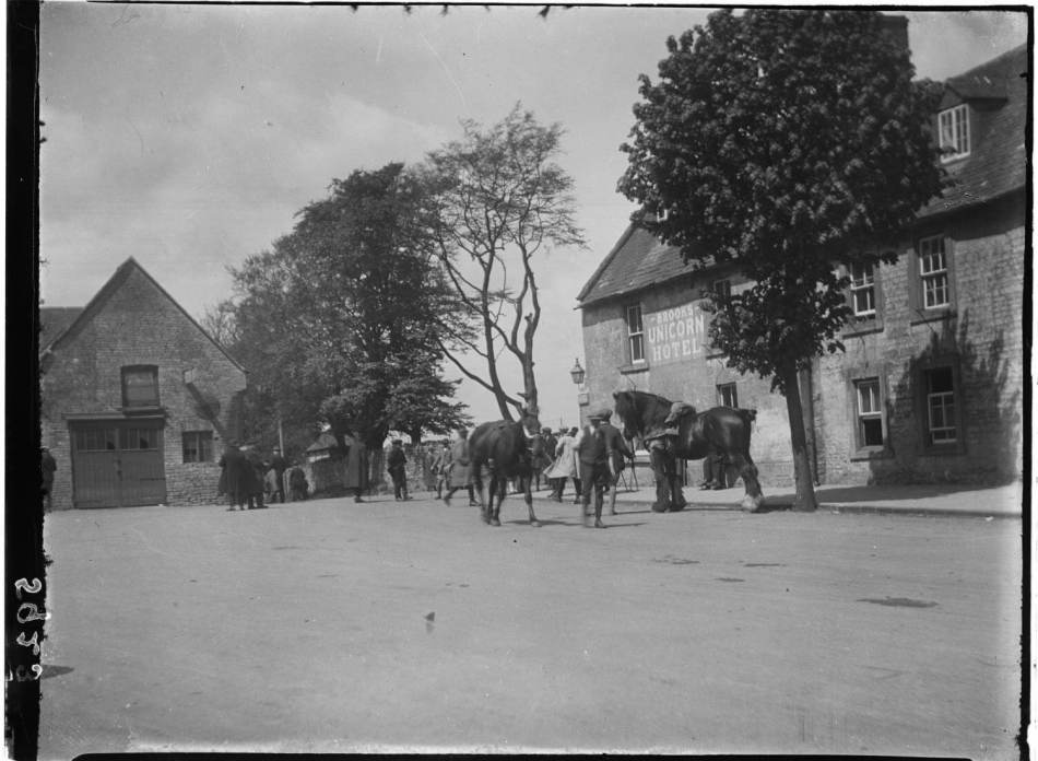 A black and white photograph of men with horses outside a hotel. 