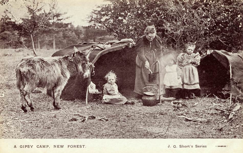 A black and white photograph of a family of Gypsies cooking over a fire. A donkey stands to the side. 