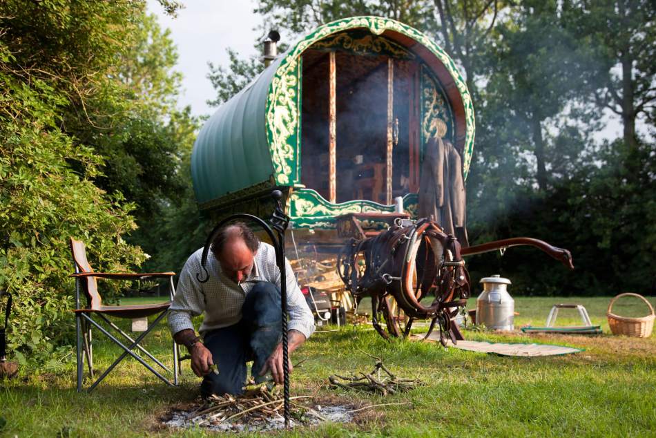 A photograph of a man tending a camp fire. In the background is a traditional Gypsy caravan. 