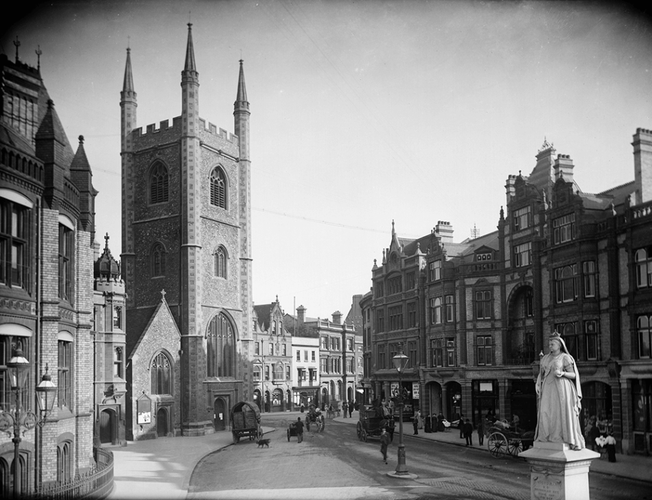 A black and white photograph of a high street with a Church tower in the background and a statue of Queen Victoria in the foreground.