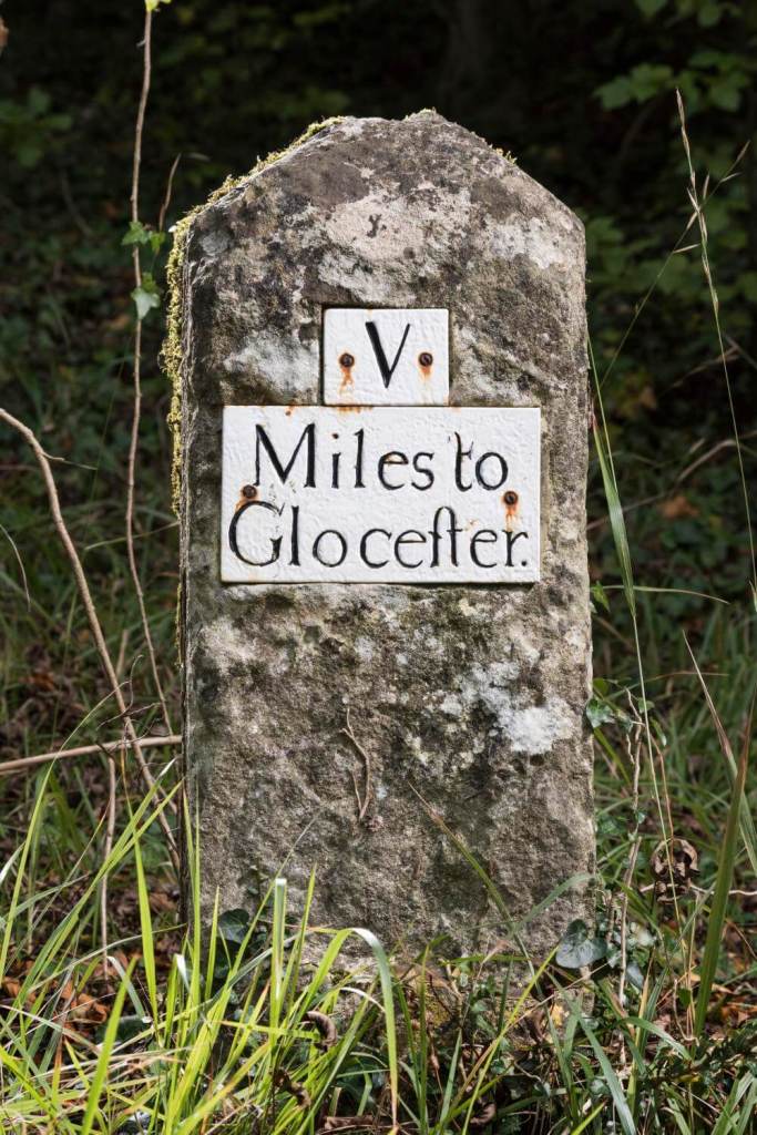 A photograph of a small milestone surrounded by grass with the wording 'V Miles to Glocester' inscribed on it.