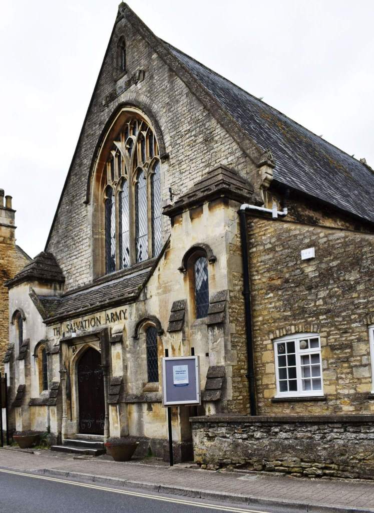 Large building constructed of Cotswold stone in Gothic style. There is a single-storey entrance hall, flanked by low towers, and a later extension to the right.