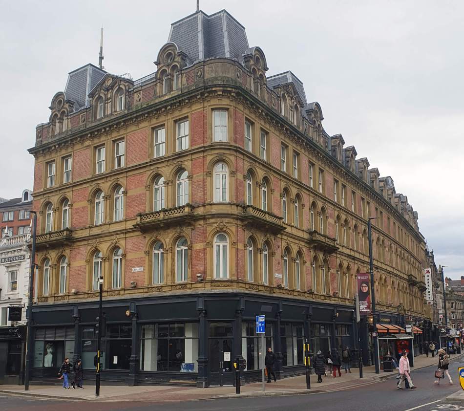 Impressive long red brick building of 4 storeys plus attic. There are shops on the ground floor, and a Mansard roof.