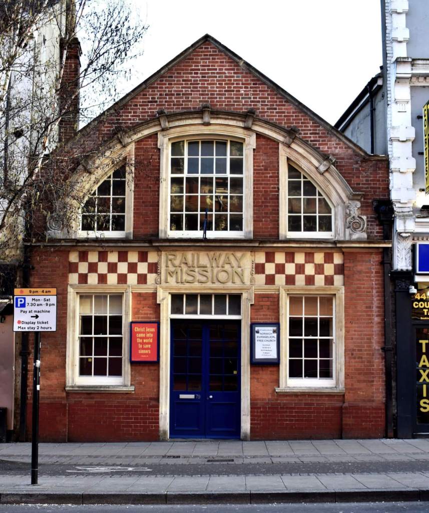 A gable end-on building in red brick with stone dressings. “Railway Mission” appears above the central door, on a chequered band beneath the string course.