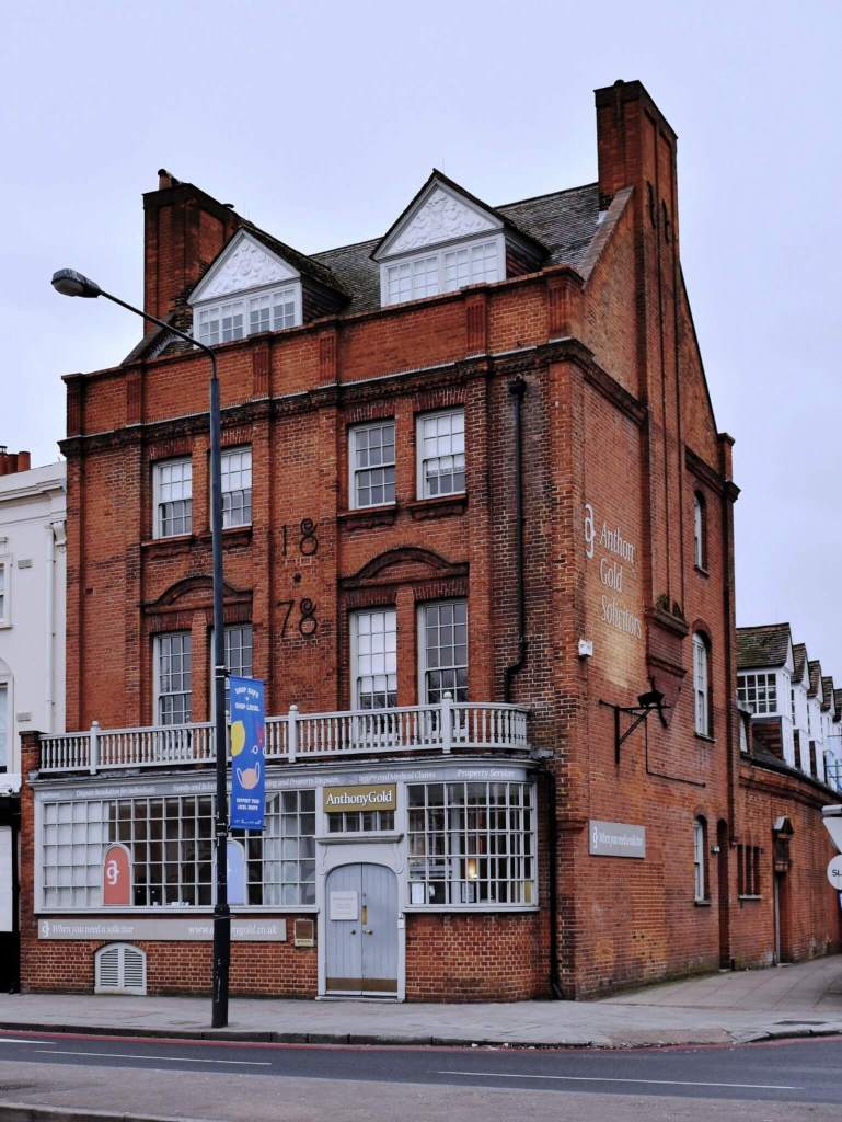 Red brick building of three storeys with dormers above, in ‘Queen Anne’ style, bearing the date ‘1878’. The glazed ground floor extends over the pavement.