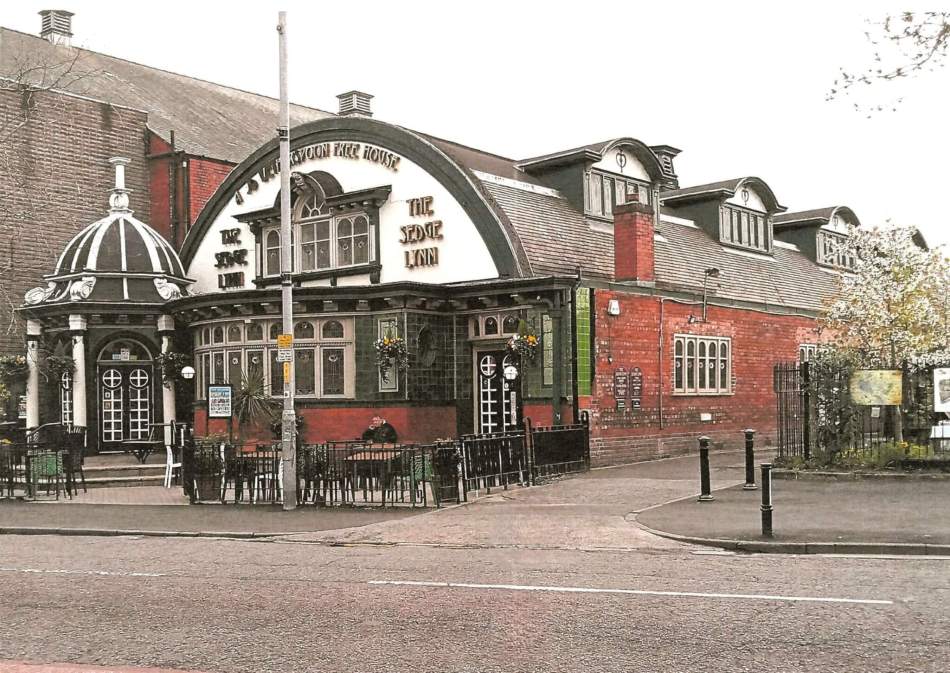 A barrel-roofed building with dormer windows, end-on to the street. The façade has much green tiling, and there is an elaborate domed pavilion to the left. 