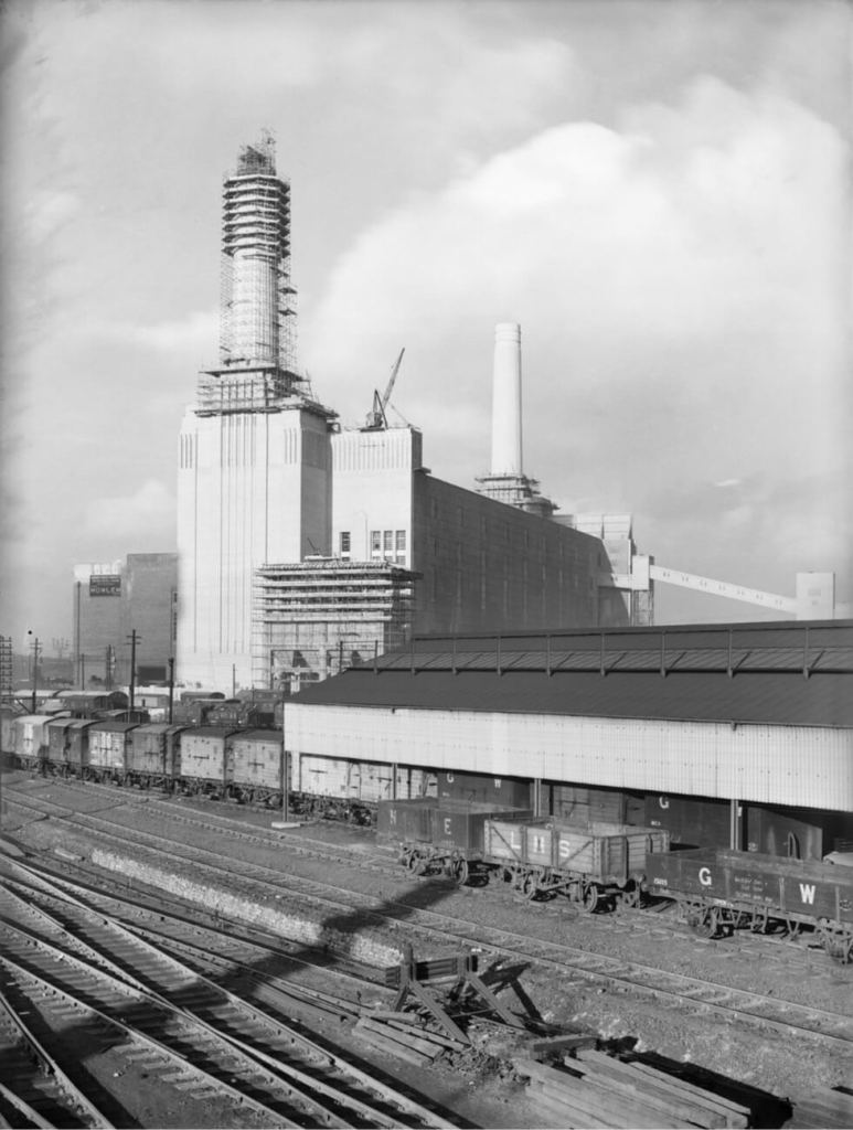 Black and white photo of a power station with 2 chimneys and railway tracks and wagons in the foreground.