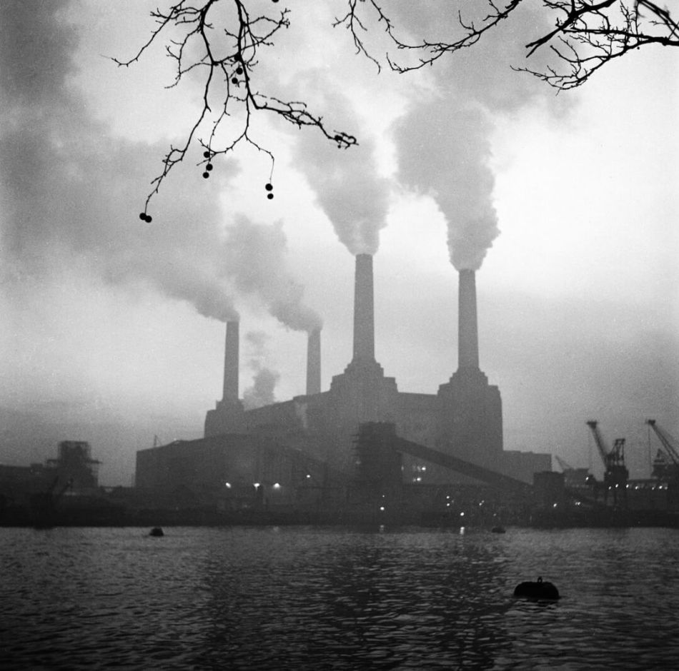 Industrial landscape with huge structure belching steam from 4 chimneys, viewed across a body of water.
